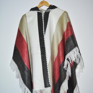 Puede incluir: Un poncho colorido con capucha, con rayas rojas, negras, beige y blancas. El poncho tiene un dobladillo con flecos y está colgado de una percha de madera. El diseño incluye una franja negra central y un borde blanco.