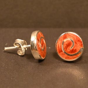 Puede incluir: Un par de pendientes de plata con diseño circular. Cada pendiente presenta un centro naranja vibrante con un patrón en espiral y un borde plateado. Los pendientes se muestran sobre un fondo oscuro.