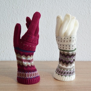 Può includere: Due sculture decorative a forma di mano in maglia. Una è rosso intenso con motivi geometrici blu, bianchi e beige. L'altra è bianco sporco con motivi geometrici verdi, marroni e viola. Entrambe sono su una superficie di legno.