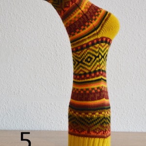 May include: A single, patterned sock in shades of yellow, orange, brown, and black. The sock has a yellow toe and cuff, with a repeating geometric design. The number "5" is visible in the image. The sock is displayed on a wooden surface.