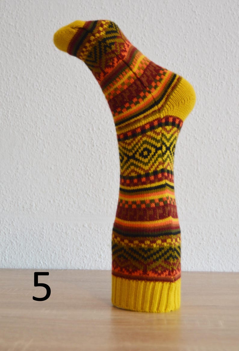 May include: A single, patterned sock in shades of yellow, orange, brown, and black. The sock has a yellow toe and cuff, with a repeating geometric design. The number "5" is visible in the image. The sock is displayed on a wooden surface.