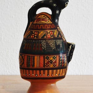 May include: A hand-painted ceramic jug with a black handle and spout. The jug features intricate geometric patterns in orange, red, green, and black. The top of the jug is adorned with a black jaguar head.