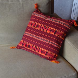 Puede incluir: Una almohada roja y naranja a rayas con un patrón geométrico y borlas. La almohada está en un sofá gris claro.