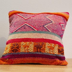 May include: A square decorative pillow with a vibrant, woven design. The pillow features a mix of colors, including pink, red, orange, and teal, with geometric and striped patterns. The pillow is resting on a wooden surface.