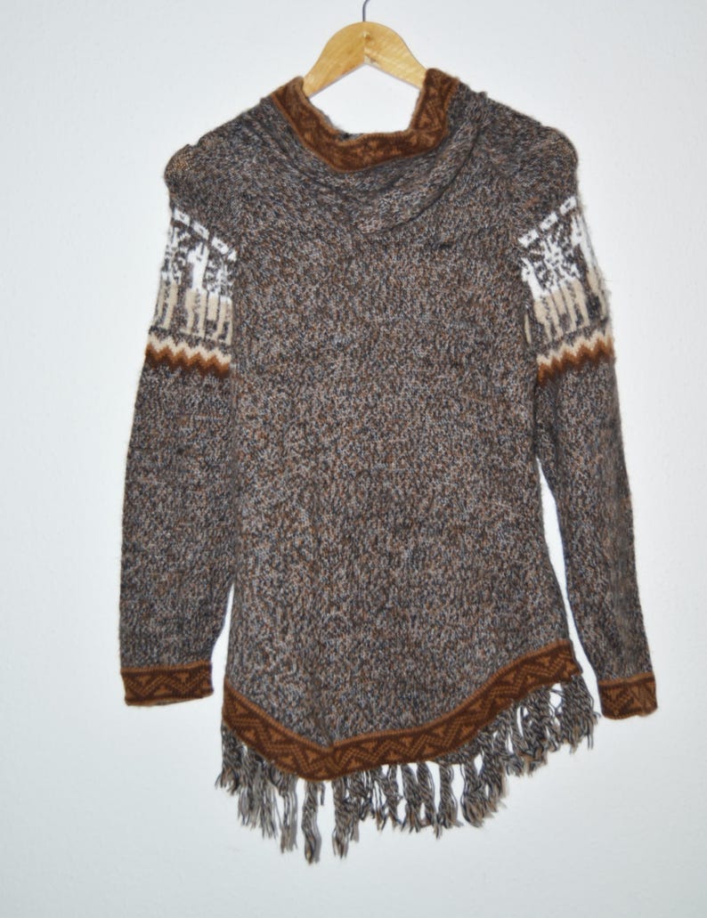 May include: A brown and gray knit sweater with a hood and fringe trim. The sweater features white and brown patterned accents on the sleeves and a brown trim along the bottom edge. The sweater is hanging on a wooden hanger.