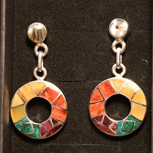 Puede incluir: Un par de pendientes colgantes de plata. Cada pendiente presenta un colgante circular con un diseño de mosaico en tonos naranja, amarillo, verde y morado. Los pendientes tienen una pequeña parte superior redonda con un acabado nacarado.