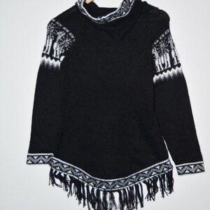 May include: Black hooded poncho with white geometric and animal pattern accents. The poncho features a fringed hem and is displayed on a wooden hanger. The sleeves and hood are trimmed with white patterns.