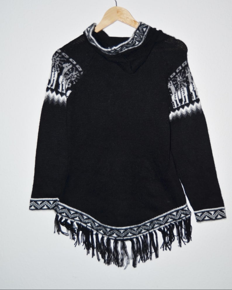 May include: Black hooded poncho with white geometric and animal pattern accents. The poncho features a fringed hem and is displayed on a wooden hanger. The sleeves and hood are trimmed with white patterns.
