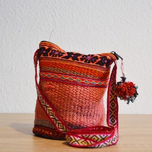 Peut inclure: Sac à bandoulière tissé aux tons orange, rouge et marron, avec des motifs géométriques complexes sur la sangle et la bordure. Une petite bourse assortie est posée à côté du sac. Un pompon rouge est suspendu à la fermeture éclair.