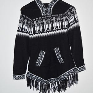 May include: Black hooded poncho with white and gray geometric patterns and fringe trim. The poncho features a front pocket and a hood with a decorative design. The design includes images of llamas.