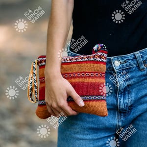 Puede incluir: Un bolso de mano rectangular tejido con un vibrante patrón de rayas rojas, naranjas y azules. El bolso de mano tiene una correa a juego y una borla decorativa. El fondo está borroso, lo que sugiere un entorno al aire libre.