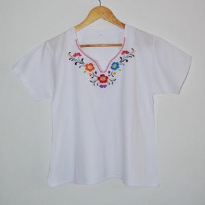 Peut inclure: T-shirt blanc à manches courtes avec un col en V. Le col est orné de broderies florales colorées rouges, oranges, bleues et violettes. Le t-shirt est suspendu à un cintre en bois.
