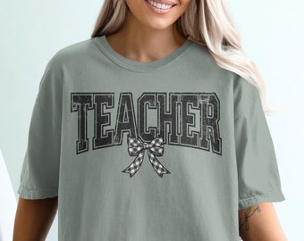 Peut inclure: Un t-shirt vert sauge avec le mot "TEACHER" en lettres noires en bloc avec un nœud à carreaux noir et blanc.
