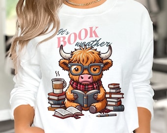 Peut inclure: Sweat-shirt blanc avec une vache Highland brune portant des lunettes et lisant un livre. Le texte "It's Book Weather" est imprimé sur le t-shirt.