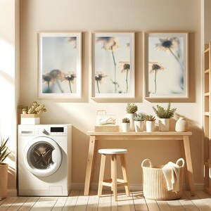 Puede incluir: Una lavadora blanca con una puerta de carga frontal, una mesa de madera con un taburete blanco, una cesta de mimbre y tres impresiones enmarcadas de flores blancas en una pared de color beige claro.