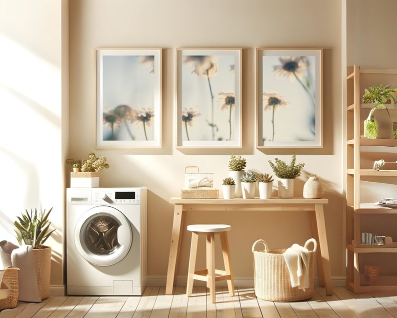 Puede incluir: Una lavadora blanca con una puerta de carga frontal, una mesa de madera con un taburete blanco, una cesta de mimbre y tres impresiones enmarcadas de flores blancas en una pared de color beige claro.