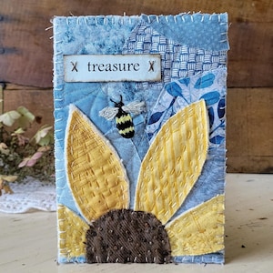 May include: A rectangular fabric art piece featuring a sunflower with yellow petals and a dark brown center, a small embroidered bee, and a white label with the text "treasure" flanked by two "X" symbols, all set against a blue patchwork background.