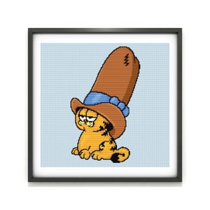 May include: A framed cross-stitch featuring Garfield the cat wearing a tall brown hat with a blue band. The orange cat is seated with a grumpy expression. The background is a light blue grid pattern.