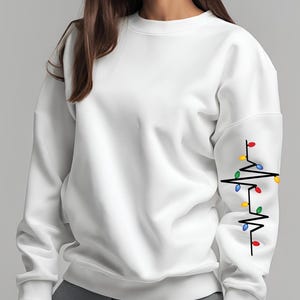 May include: White sweatshirt with a crew neck. The left sleeve features a black heartbeat line design with colorful Christmas lights. The lights are red, yellow, green, and blue.