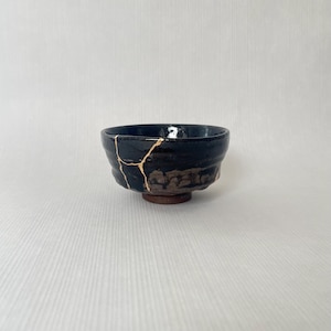 May include: A black ceramic bowl with gold kintsugi repair lines. The bowl has a rough, textured exterior and sits on a small, dark brown base. The interior of the bowl is smooth and glossy.
