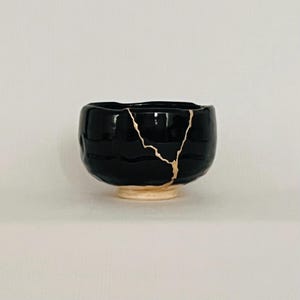 May include: A black ceramic bowl with a gold crack pattern, on a beige base. The bowl has a glossy finish and a textured exterior. This piece showcases the Japanese art of Kintsugi, a pottery repair method.
