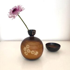 May include: A wooden vase with a dark brown finish and a carved design of white flowers. A single pink flower is in the vase. A small, dark brown wooden bowl is next to the vase.