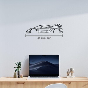 Puede incluir: Una pieza de arte mural de metal negro que representa un coche deportivo. La obra de arte es un dibujo lineal de un coche con aler&oacute;n, que mide 40 cm de largo. El coche est&aacute; montado en una pared blanca sobre un escritorio.