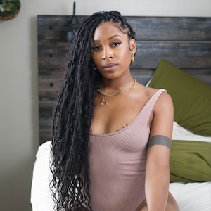 May include: A woman with long, dark, curly hair is wearing a sleeveless, ribbed, mauve bodysuit. She is sitting on a white bed with a green pillow behind her. She is wearing gold hoop earrings and a gold necklace.