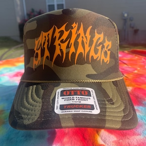 Può includere: Un cappellino da camionista mimetico con la scritta "SPRING" in arancione, con lettere stilizzate sul davanti. Il cappello ha una visiera curva e una chiusura a scatto. L'etichetta recita "OTTO World Famous Foam Front Trucker".