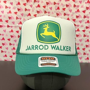 Op de afbeelding: Een witte en groene trucker-pet met een groen John Deere-logo en de tekst "Jarrod Walker" op de voorkant. De pet heeft een groen mesh-achterkant en een zwarte drukknopsluiting.