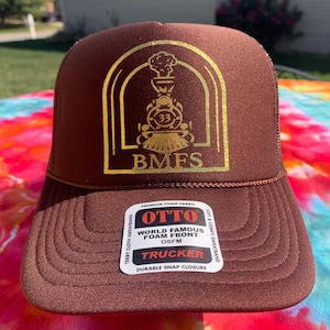 Może przedstawiać: Brązowa czapka typu trucker ze złotą grafiką pociągu i literami "BMFS". Czapka ma zakrzywiony daszek i siatkową tylną część. Czapka ma etykietę "OTTO" z napisem "World Famous Foam Front OSFM Trucker".