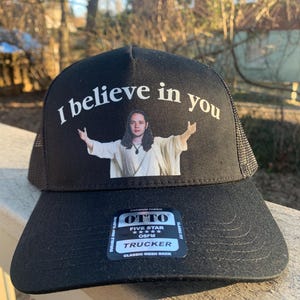 May include: Black trucker hat with a mesh back. The hat has a white graphic that says "I believe in you" and a picture of a person with their arms outstretched. The hat also has a black label with white text that says "OTTO FIVE STAR TRUCKER CLASSIC MESH BACK CLASSIC FIT".