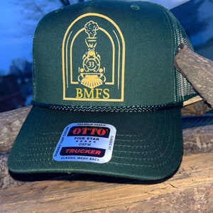 Op de afbeelding: Een groene truckercap met een geborduurd logo in goud van een trein met de tekst "BMFS" eronder. De pet heeft een mesh-rug en een label met de tekst "OTTO FIVE STAR OSFM TRUCKER CLASSIC MESH BACK".