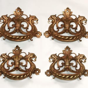 May include: Four antique bronze-colored drawer pulls. Each pull features a decorative design with a central floral element, flanked by two stylized dragon-like figures. The pulls have a curved handle and ornate detailing, suitable for furniture restoration.