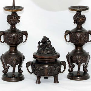 May include: Three antique bronze decorative pieces. Two tall candle holders with ornate dragon and floral designs, and a central incense burner with a detailed lid. The pieces are dark brown with intricate carvings.