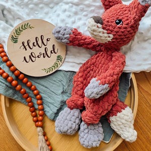 Puede incluir: Un peluche de zorro tejido a mano, rojo y gris, con hocico y cola blancos. Un letrero de madera con las palabras "Hello World" está al lado del zorro. Una bandeja de madera contiene el zorro, el letrero y un hilo de cuentas naranjas.