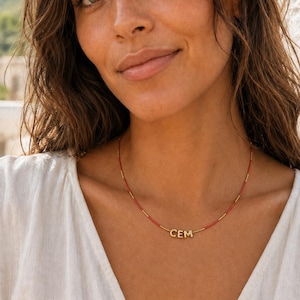 Può includere: Una collana delicata caratterizzata da un filo sottile di piccole perline rosse intervallate da minuscoli distanziatori metallici dorati. Il centro della collana mostra tre ciondoli a forma di lettera dorata che compongono la parola CEM.