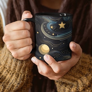 May include: A black ceramic mug with a galaxy design featuring gold stars, a crescent moon, and swirling lines on a dark blue background.