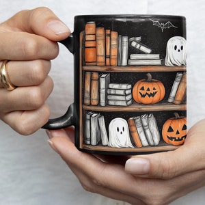 Puede incluir: Taza de cerámica negra con un diseño de temática de Halloween. La taza presenta una estantería con libros, calabazas y fantasmas.