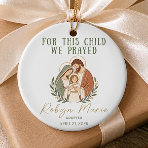 Op de afbeelding: Een rond wit keramisch ornament met een gouden koord en een crèmekleurig lint. Het ontwerp bevat een familie-illustratie, de tekst "FOR THIS CHILD WE PRAYED", de naam "Robyn Marie", het woord "ADOPTED" en "APRIL 27, 2026".