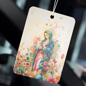 以下が含まれることがあります： 花に囲まれた聖母マリアのカラフルな画像が描かれた車の芳香剤。画像は薄い黄色の色合いの白い背景に描かれています。