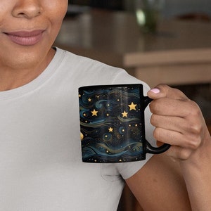 May include: A person holding a black coffee mug with a gold and blue starry night design.