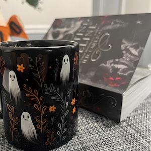 Black ceramic coffee mug featuring a whimsical Halloween design with cute white ghosts surrounded by orange, yellow, and gray autumn foliage and flowers on a dark background. The mug has a glossy finish and a large handle for comfortable use. Available in 11 oz or 15 oz sizes, this festive fall mug is both microwave safe and dishwasher safe, making it a practical yet fun seasonal drinkware choice for coffee, tea, or hot cocoa. (11 oz shown)