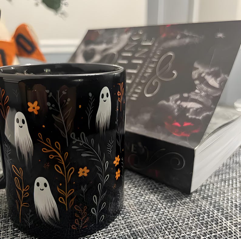 Black ceramic coffee mug featuring a whimsical Halloween design with cute white ghosts surrounded by orange, yellow, and gray autumn foliage and flowers on a dark background. The mug has a glossy finish and a large handle for comfortable use. Available in 11 oz or 15 oz sizes, this festive fall mug is both microwave safe and dishwasher safe, making it a practical yet fun seasonal drinkware choice for coffee, tea, or hot cocoa. (11 oz shown)
