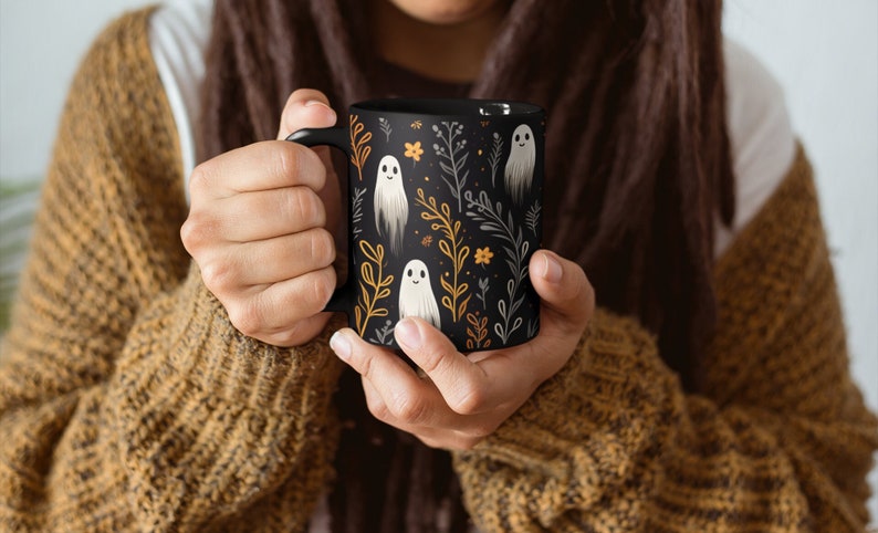 Black ceramic coffee mug featuring a whimsical Halloween design with cute white ghosts surrounded by orange, yellow, and gray autumn foliage and flowers on a dark background. The mug has a glossy finish and a large handle for comfortable use. Available in 11 oz or 15 oz sizes, this festive fall mug is both microwave safe and dishwasher safe, making it a practical yet fun seasonal drinkware choice for coffee, tea, or hot cocoa.