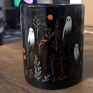 Black ceramic coffee mug featuring a whimsical Halloween design with cute white ghosts surrounded by orange, yellow, and gray autumn foliage and flowers on a dark background. The mug has a glossy finish and a large handle for comfortable use. Available in 11 oz or 15 oz sizes, this festive fall mug is both microwave safe and dishwasher safe, making it a practical yet fun seasonal drinkware choice for coffee, tea, or hot cocoa. (11 oz shown)