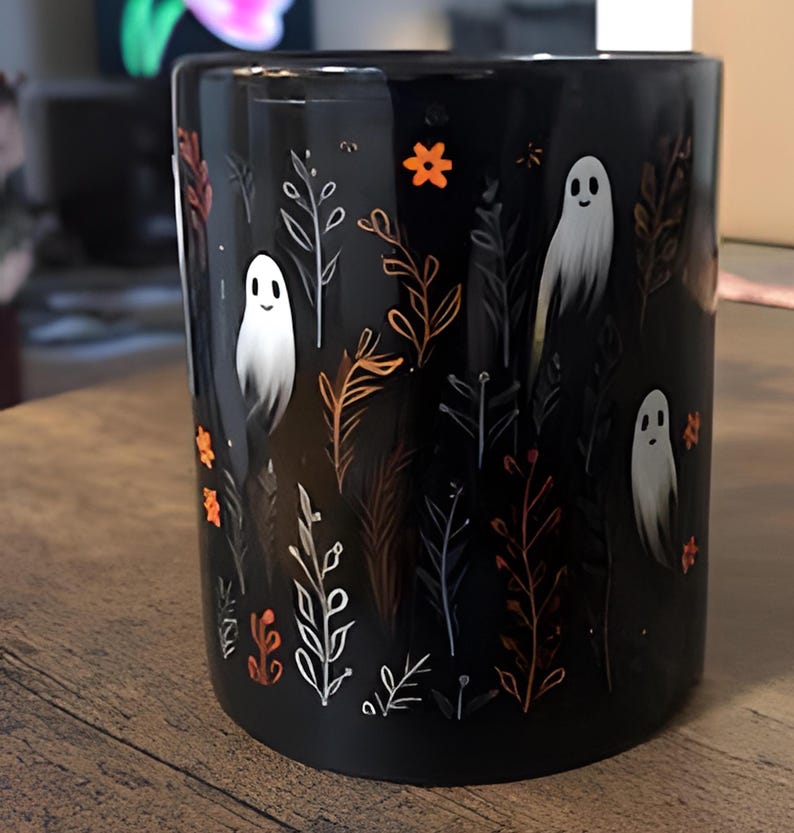 Black ceramic coffee mug featuring a whimsical Halloween design with cute white ghosts surrounded by orange, yellow, and gray autumn foliage and flowers on a dark background. The mug has a glossy finish and a large handle for comfortable use. Available in 11 oz or 15 oz sizes, this festive fall mug is both microwave safe and dishwasher safe, making it a practical yet fun seasonal drinkware choice for coffee, tea, or hot cocoa. (11 oz shown)