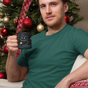 May include: A man wearing a teal green t-shirt is holding a black mug with a gold and blue starry night design. He is sitting in front of a Christmas tree decorated with red and gold ornaments.