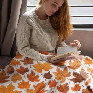 Op de afbeelding: Een wit deken met een patroon van oranje en bruine herfstbladeren. Een persoon zit op het deken en leest een boek.