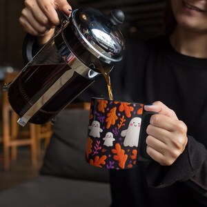 May include: A black coffee mug with a Halloween theme. The mug features white ghost illustrations and orange leaf designs on a black background. Coffee is being poured from a French press into the mug.
